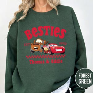May include: Forest green sweatshirt featuring the word "Besties" in red, with cartoon car graphics. The design includes the text "Est. 1996" and the names "Thomas & Eddie" in red.