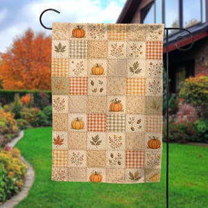 May include: A rectangular garden flag featuring a patchwork quilt design with alternating squares of orange pumpkins, brown acorns, autumn leaves, and tan gingham patterns in shades of orange, beige, and muted green on a textured fabric background.