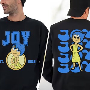 May include: Black sweatshirt featuring the word "JOY" in blue block letters with a cartoon character illustration. The back of the sweatshirt has the word "JOY" repeated in a pattern with the same character.