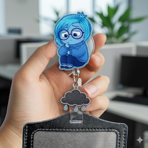 May include: A blue and white cartoon character badge reel featuring a sad-looking character with blue hair and glasses. The badge reel has a black cloud charm with raindrops and a black ID holder.