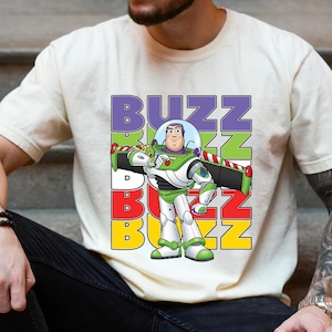Puede incluir: Camiseta color crema con un gr&aacute;fico de Buzz Lightyear de Toy Story. El dise&ntilde;o incluye la palabra "BUZZ" en letras apiladas y coloridas. Buzz se representa con su traje espacial verde y blanco caracter&iacute;stico.