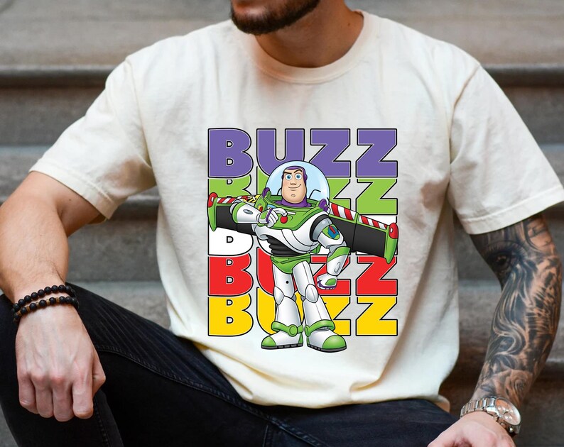 Puede incluir: Camiseta color crema con un gr&aacute;fico de Buzz Lightyear de Toy Story. El dise&ntilde;o incluye la palabra "BUZZ" en letras apiladas y coloridas. Buzz se representa con su traje espacial verde y blanco caracter&iacute;stico.
