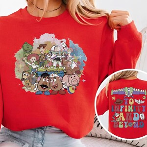May include: A red sweatshirt featuring a colorful graphic of characters from the animated film "Toy Story." The front of the sweatshirt displays a group of characters, while the back has the phrase "To Infinity and Beyond."