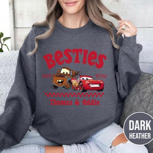 May include: Dark heather gray sweatshirt featuring the word "Besties" in red, with cartoon images of Mater and Lightning McQueen. The text "1996" and the names "Thomas & Eddie" are also in red. The sweatshirt has a crew neck.