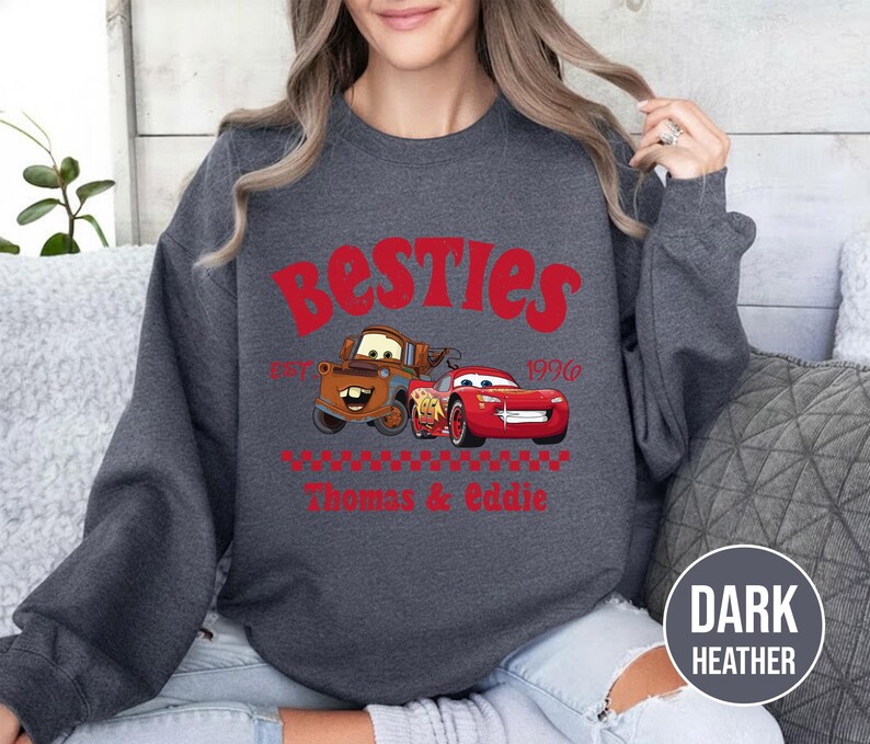 May include: Dark heather gray sweatshirt featuring the word "Besties" in red, with cartoon images of Mater and Lightning McQueen. The text "1996" and the names "Thomas & Eddie" are also in red. The sweatshirt has a crew neck.