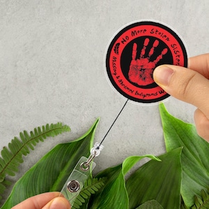 May include: A round badge with the text "No More Stolen Sisters" and a red handprint on a black background. The badge is attached to a retractable reel with a clear plastic clip. The image is set against a backdrop of green leaves.