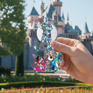 May include: A set of three character-shaped acrylic charms featuring Hades and his henchmen from the animated film. The charms are colourful with geometric patterns and are held in front of a castle.
