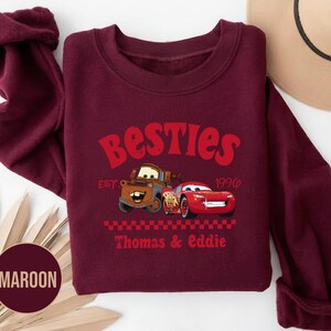 May include: Maroon sweatshirt featuring the text "Besties" in red, with cartoon car graphics. The design includes the text "EST. 1996" and "Thomas & Eddie". The sweatshirt has a crew neck and long sleeves.