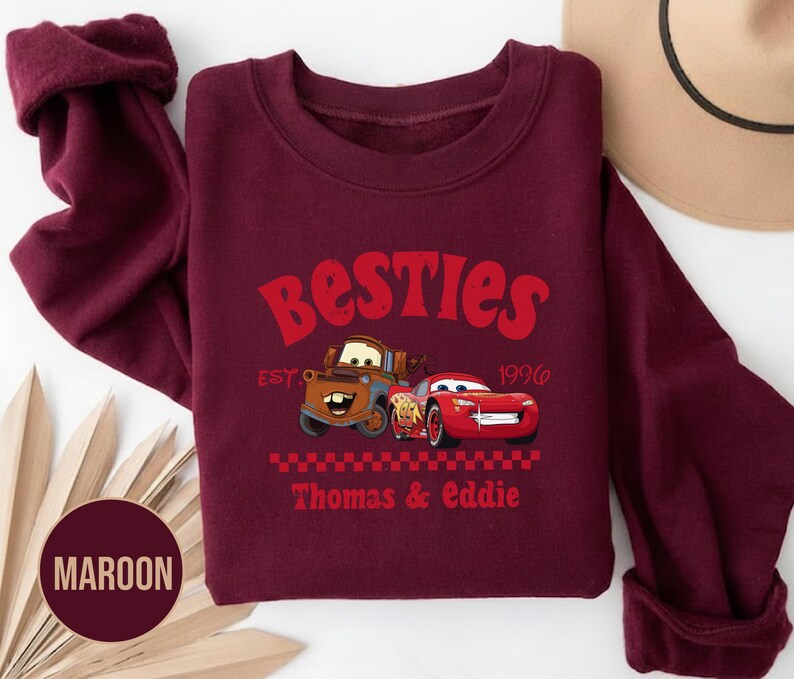 May include: Maroon sweatshirt featuring the text "Besties" in red, with cartoon car graphics. The design includes the text "EST. 1996" and "Thomas & Eddie". The sweatshirt has a crew neck and long sleeves.