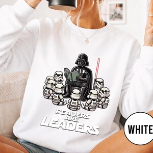 K&ouml;nnte beinhalten: Wei&szlig;es Sweatshirt mit einer Cartoon-Illustration von Darth Vader, der einer Gruppe von Sturmtruppen vorliest. Der Text "Readers are Leaders" ist unter der Illustration aufgedruckt. Ein rotes Lichtschwert ist ebenfalls zu sehen.
