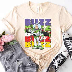 Puede incluir: Camiseta color crema con un dise&ntilde;o de Buzz Lightyear y la palabra "BUZZ" en bloques coloridos. El dise&ntilde;o incluye una imagen de dibujos animados de Buzz con su traje espacial. La camiseta se combina con vaqueros grises y zapatillas negras.