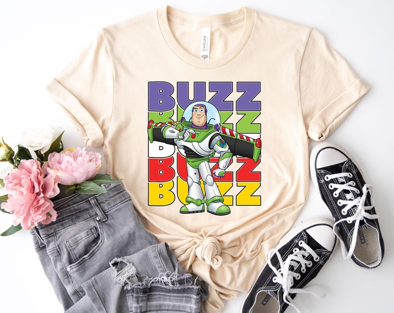 Puede incluir: Camiseta color crema con un dise&ntilde;o de Buzz Lightyear y la palabra "BUZZ" en bloques coloridos. El dise&ntilde;o incluye una imagen de dibujos animados de Buzz con su traje espacial. La camiseta se combina con vaqueros grises y zapatillas negras.