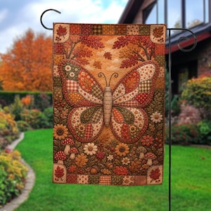 May include: A rectangular garden flag featuring a central butterfly with patchwork-style wings in shades of orange, brown, and green. The design includes autumn leaves, mushrooms, sunflowers, and acorns, all framed by a checkered border pattern.