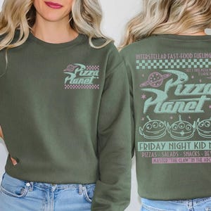 May include: Olive green sweatshirt with the Pizza Planet logo on the front and back. The front features the logo in pink and white. The back has the Pizza Planet logo, aliens, and text in pink and teal.