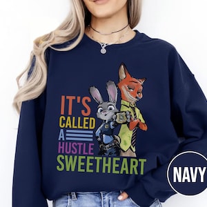 Puede incluir: Sudadera azul marino con un gr&aacute;fico colorido de Judy Hopps y Nick Wilde de Zootopia, con el texto "IT'S CALLED A HUSTLE SWEETHEART". La sudadera tiene cuello redondo y mangas largas.