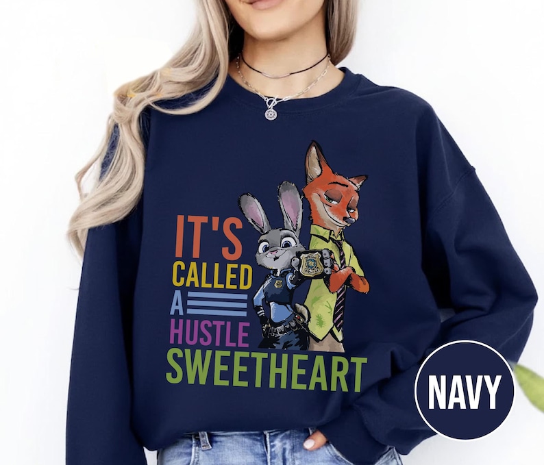 Puede incluir: Sudadera azul marino con un gr&aacute;fico colorido de Judy Hopps y Nick Wilde de Zootopia, con el texto "IT'S CALLED A HUSTLE SWEETHEART". La sudadera tiene cuello redondo y mangas largas.
