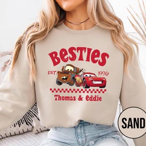 May include: Sand-colored sweatshirt featuring a graphic of two cartoon cars and the text "Besties Est. 1996 Thomas & Eddie" in red. The design includes a brown tow truck and a red race car.