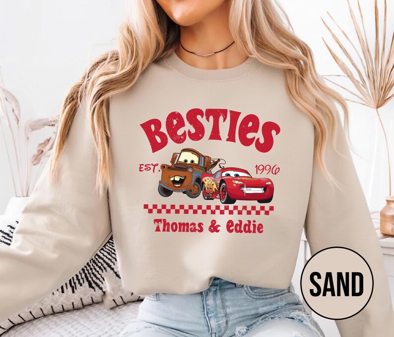 May include: Sand-colored sweatshirt featuring a graphic of two cartoon cars and the text "Besties Est. 1996 Thomas & Eddie" in red. The design includes a brown tow truck and a red race car.
