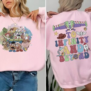 May include: A light pink sweatshirt featuring a colorful graphic of Toy Story characters on the front. The back of the sweatshirt has a graphic of Buzz Lightyear's ship and the phrase "To Infinity and Beyond" in a retro font.