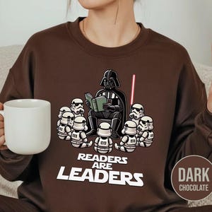 K&ouml;nnte beinhalten: Dunkel schokoladenbraunes Sweatshirt mit einer Cartoon-Illustration von Darth Vader, der Stormtroopern vorliest. Der Text "Readers Are Leaders" ist darunter gedruckt. Ein rotes Lichtschwert ist ebenfalls zu sehen.