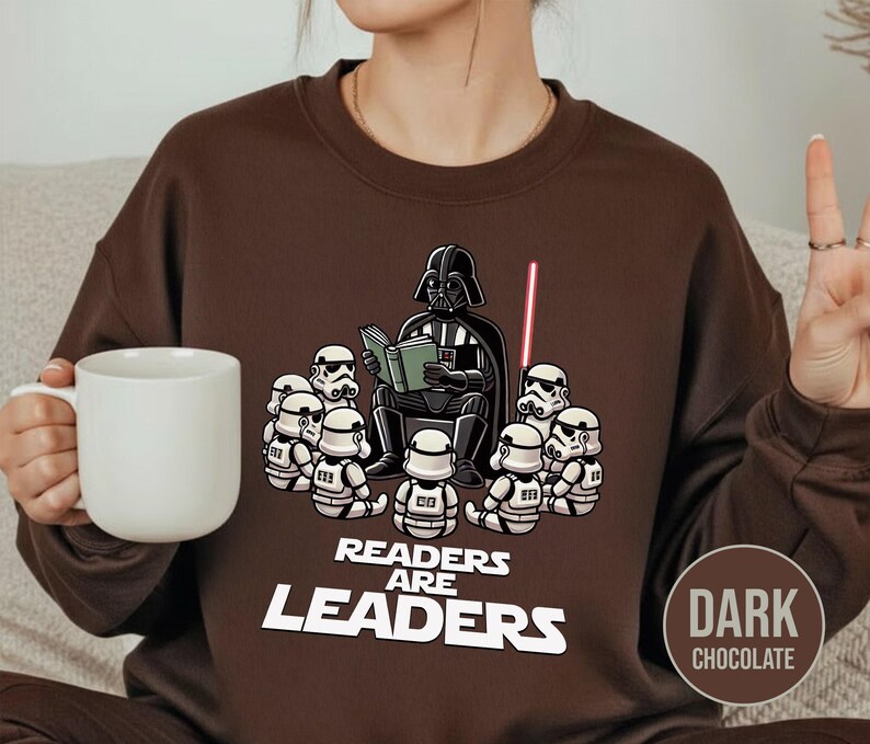 K&ouml;nnte beinhalten: Dunkel schokoladenbraunes Sweatshirt mit einer Cartoon-Illustration von Darth Vader, der Stormtroopern vorliest. Der Text "Readers Are Leaders" ist darunter gedruckt. Ein rotes Lichtschwert ist ebenfalls zu sehen.