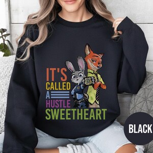 Puede incluir: Sudadera negra con un gr&aacute;fico colorido de Judy Hopps y Nick Wilde de Zootopia, con el texto "IT'S CALLED A HUSTLE SWEETHEART". La sudadera tiene cuello redondo y mangas largas.