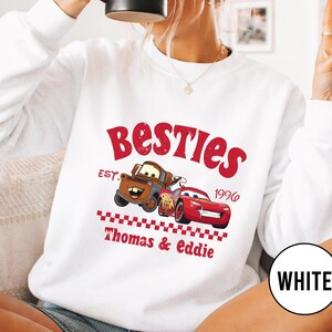 May include: White sweatshirt featuring a red graphic with the word "Besties" and the year "1996". The graphic includes cartoon images of two cars. The text "Thomas & Eddie" is also printed on the sweatshirt.