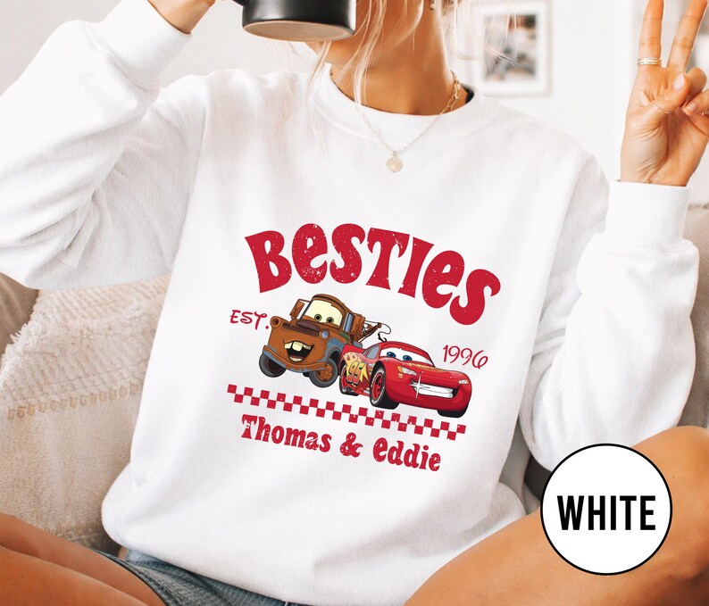 May include: White sweatshirt featuring a red graphic with the word "Besties" and the year "1996". The graphic includes cartoon images of two cars. The text "Thomas & Eddie" is also printed on the sweatshirt.