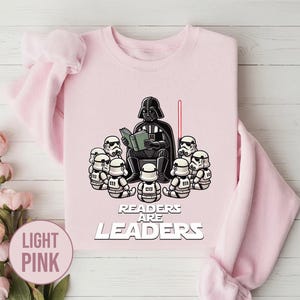 K&ouml;nnte beinhalten: Hellrosa Sweatshirt mit einer Grafik von Darth Vader, der Stormtroopern vorliest. Der Text "Readers Are Leaders" ist unter der Illustration aufgedruckt. Ein rotes Lichtschwert ist ebenfalls im Bild.