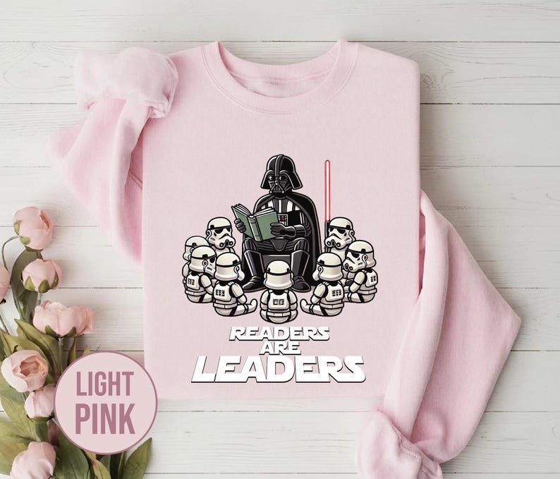 K&ouml;nnte beinhalten: Hellrosa Sweatshirt mit einer Grafik von Darth Vader, der Stormtroopern vorliest. Der Text "Readers Are Leaders" ist unter der Illustration aufgedruckt. Ein rotes Lichtschwert ist ebenfalls im Bild.