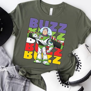 Puede incluir: Camiseta verde oliva con un gr&aacute;fico de Buzz Lightyear y la palabra "BUZZ" en letras coloridas apiladas. El personaje lleva su traje espacial verde y blanco. La camiseta se combina con botas blancas y vaqueros grises.