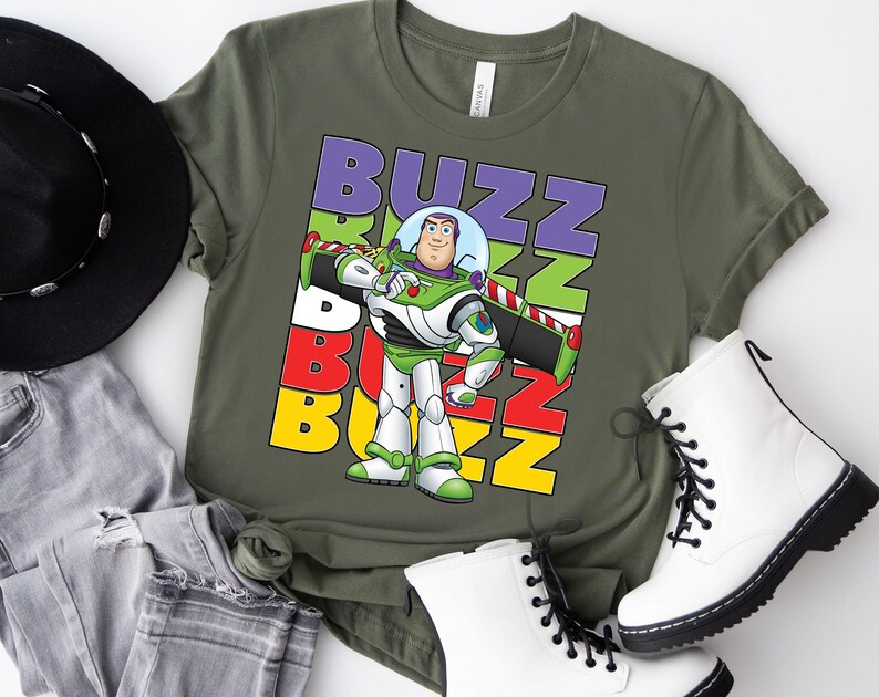 Puede incluir: Camiseta verde oliva con un gr&aacute;fico de Buzz Lightyear y la palabra "BUZZ" en letras coloridas apiladas. El personaje lleva su traje espacial verde y blanco. La camiseta se combina con botas blancas y vaqueros grises.