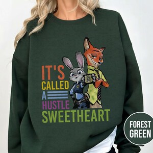 Puede incluir: Sudadera verde bosque con un dise&ntilde;o de Judy Hopps y Nick Wilde de Zootopia. El texto "IT'S CALLED A HUSTLE SWEETHEART" est&aacute; impreso en letras coloridas. La sudadera es de cuello redondo.