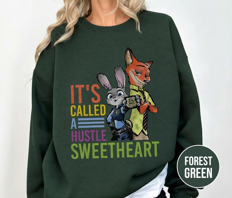 Puede incluir: Sudadera verde bosque con un dise&ntilde;o de Judy Hopps y Nick Wilde de Zootopia. El texto "IT'S CALLED A HUSTLE SWEETHEART" est&aacute; impreso en letras coloridas. La sudadera es de cuello redondo.