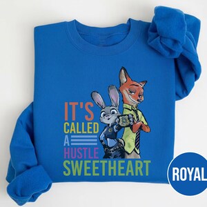 Puede incluir: Sudadera azul real con un gr&aacute;fico de un zorro y un conejo con uniformes de polic&iacute;a. El texto "IT'S CALLED A HUSTLE SWEETHEART" est&aacute; impreso en naranja, azul y verde. La sudadera tiene mangas largas y cuello redondo.