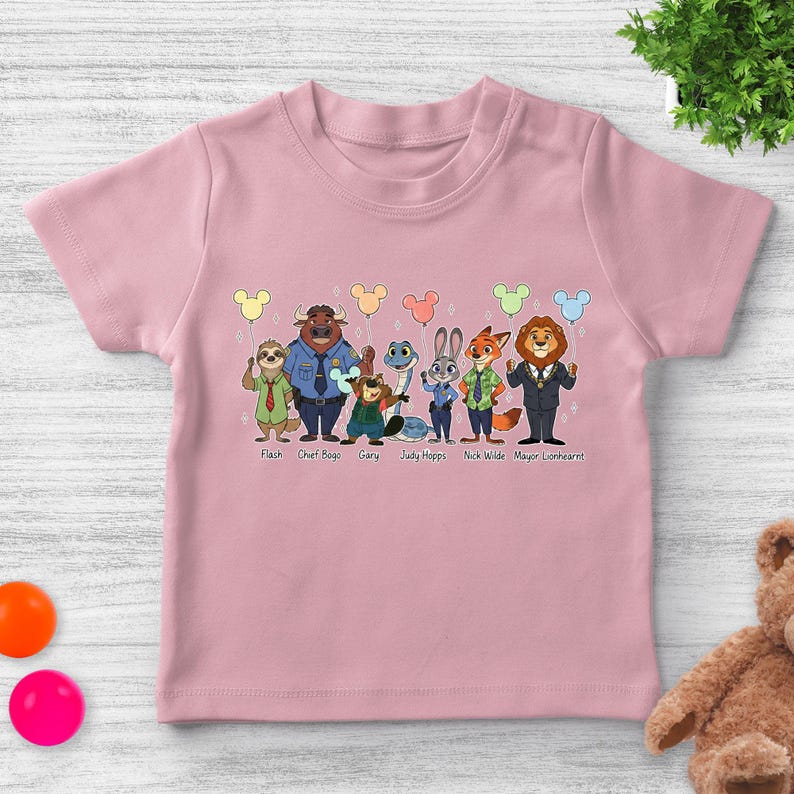 May include: Pink toddler t-shirt featuring cartoon characters from Zootopia, including Flash, Chief Bogo, and Judy Hopps. Each character holds a Mickey Mouse-shaped balloon. The names of the characters are printed below them.