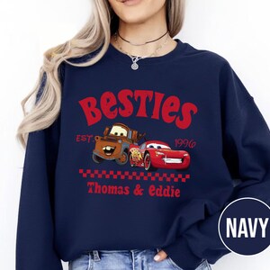 May include: Navy blue sweatshirt featuring the word "Besties" in red, with cartoon images of two cars. The sweatshirt includes the text "EST. 1996" and "Thomas & Eddie" in red. A navy blue circle with the word "NAVY" is in the bottom right corner.
