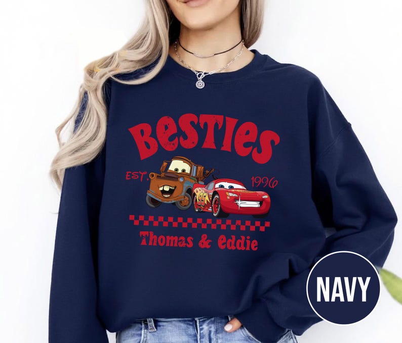 May include: Navy blue sweatshirt featuring the word "Besties" in red, with cartoon images of two cars. The sweatshirt includes the text "EST. 1996" and "Thomas & Eddie" in red. A navy blue circle with the word "NAVY" is in the bottom right corner.