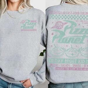 May include: A light gray sweatshirt featuring the Pizza Planet logo in mint and pink. The back of the sweatshirt has the text "Interstellar Fast-Food Fueling Station" and "Friday Night Kid Nite" with alien graphics.