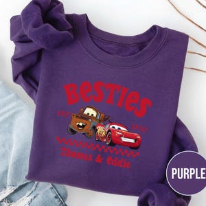 May include: A purple sweatshirt featuring the text "Besties" in red, with the image of two cartoon cars. The sweatshirt also includes the text "Est. 1996" and the names "Thomas & Eddie" in red. A purple circle with the word "PURPLE" is in the lower right corner.