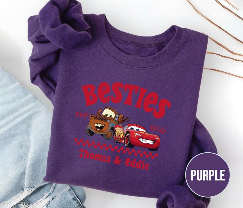 May include: A purple sweatshirt featuring the text "Besties" in red, with the image of two cartoon cars. The sweatshirt also includes the text "Est. 1996" and the names "Thomas & Eddie" in red. A purple circle with the word "PURPLE" is in the lower right corner.