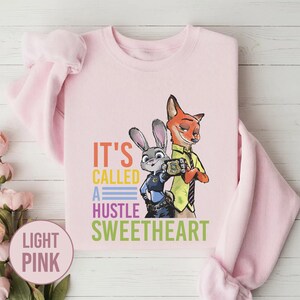 Puede incluir: Sudadera rosa claro con un dise&ntilde;o de un conejo y un zorro de Zootopia, con el texto "It's Called A Hustle Sweetheart". La sudadera es de cuello redondo.
