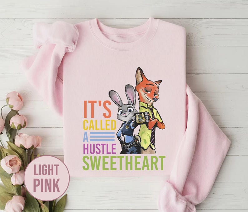 Puede incluir: Sudadera rosa claro con un dise&ntilde;o de un conejo y un zorro de Zootopia, con el texto "It's Called A Hustle Sweetheart". La sudadera es de cuello redondo.