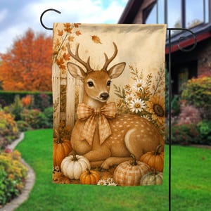 May include: A rectangular garden flag featuring a brown deer with antlers wearing a tan plaid bow. The deer is surrounded by orange and white pumpkins, white daisies, a sunflower, and a wooden fence against a beige background.
