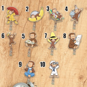 May include: A collection of ten acrylic badge reels featuring the Curious George character in various poses. The designs include George with an umbrella, on a banana, relaxing in a chair, with a birthday cake, reading a book, and wearing a sombrero.