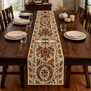 May include: A long, rectangular table runner featuring a cream-colored background with a repeating pattern of orange and red flowers, green leaves, and brown acorns. The fabric has a quilted texture and is centered on a dark wooden dining table.