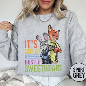 Puede incluir: Sudadera gris deportiva con un gr&aacute;fico colorido de Judy Hopps y Nick Wilde de Zootopia, con el texto "IT'S CALLED A HUSTLE SWEETHEART". La sudadera tiene cuello redondo y mangas largas.