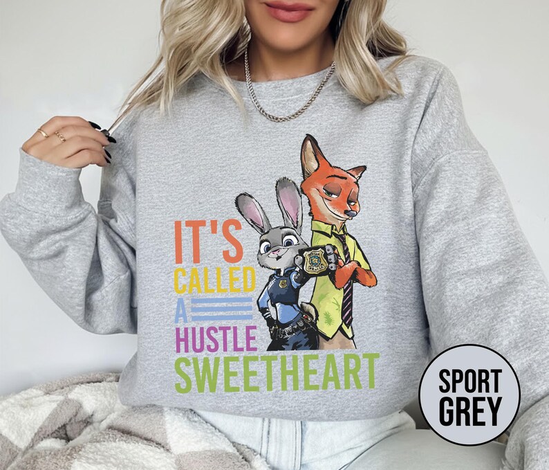 Puede incluir: Sudadera gris deportiva con un gr&aacute;fico colorido de Judy Hopps y Nick Wilde de Zootopia, con el texto "IT'S CALLED A HUSTLE SWEETHEART". La sudadera tiene cuello redondo y mangas largas.