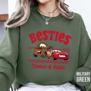 May include: Military green sweatshirt featuring the text "Besties" in red, with cartoon images of Mater and Lightning McQueen. The sweatshirt also includes the text "Est. 1996" and "Thomas & Eddie".