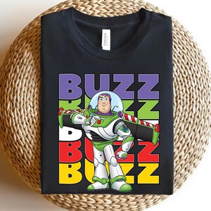 Puede incluir: Camiseta negra con un gr&aacute;fico de Buzz Lightyear de Toy Story. El dise&ntilde;o incluye la palabra "BUZZ" en letras apiladas de colores, con Buzz Lightyear en su traje espacial verde, blanco y morado. La camiseta se muestra sobre una superficie tejida.
